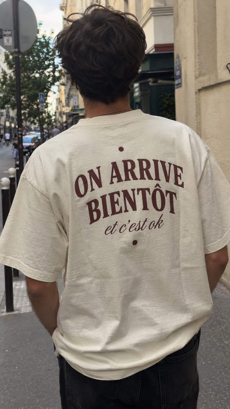 Vu de dos, un t-shirt crème imprimé en bordeaux : On arrive bientôt, et c'est ok.